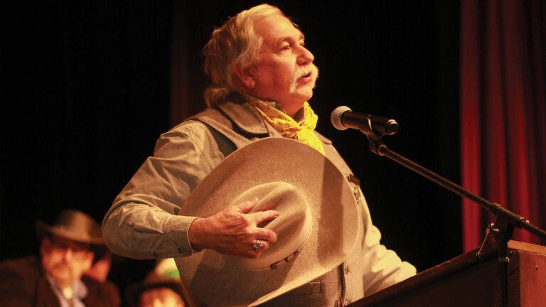man holding a cowboy hat talking at the podium at Mystery Weekend