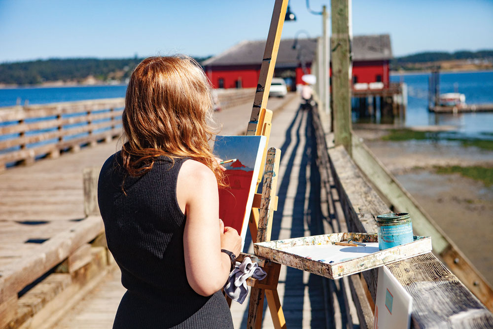 Looking behind young woman as she paints a red wharf building in the distance