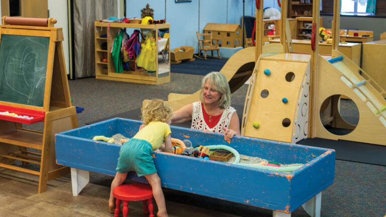 Betsy O’Neil interacts with young child in playroom sandbox