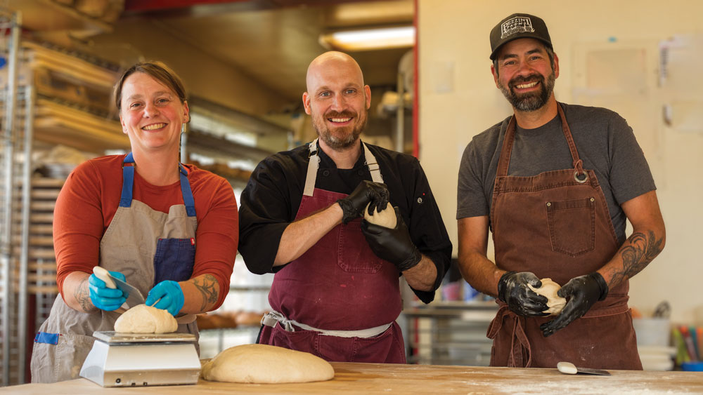 Three people, one woman on the left and two men enter and right, measuring and forming pizza dough