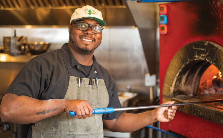 Man standing in from of wood burning oven holding pizza peel