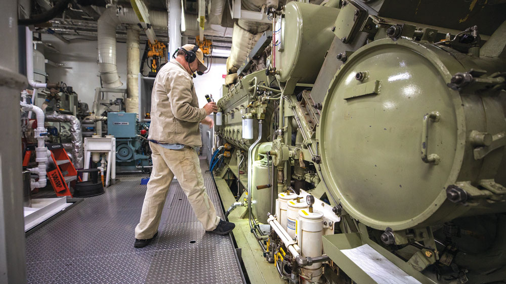 Man in tan work clothes shines flashlight into engine compartment in the engine room of a ferry