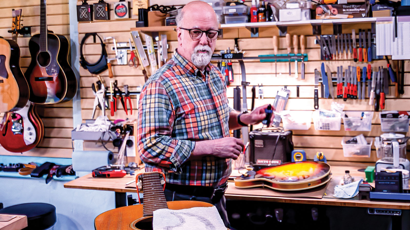 Man in a guitar workshop with tools on the wall and a guitar on his workbench.