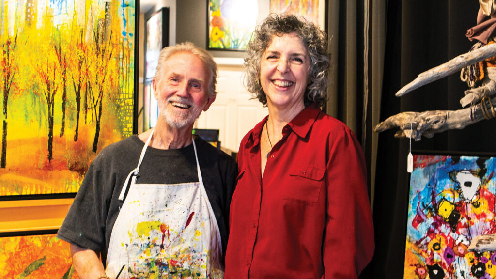 Man with painted apron and woman standing next to him, both in front of colorful paintings.
