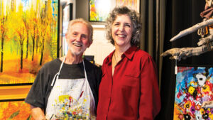Man with painted apron and woman standing next to him, both in front of colorful paintings.