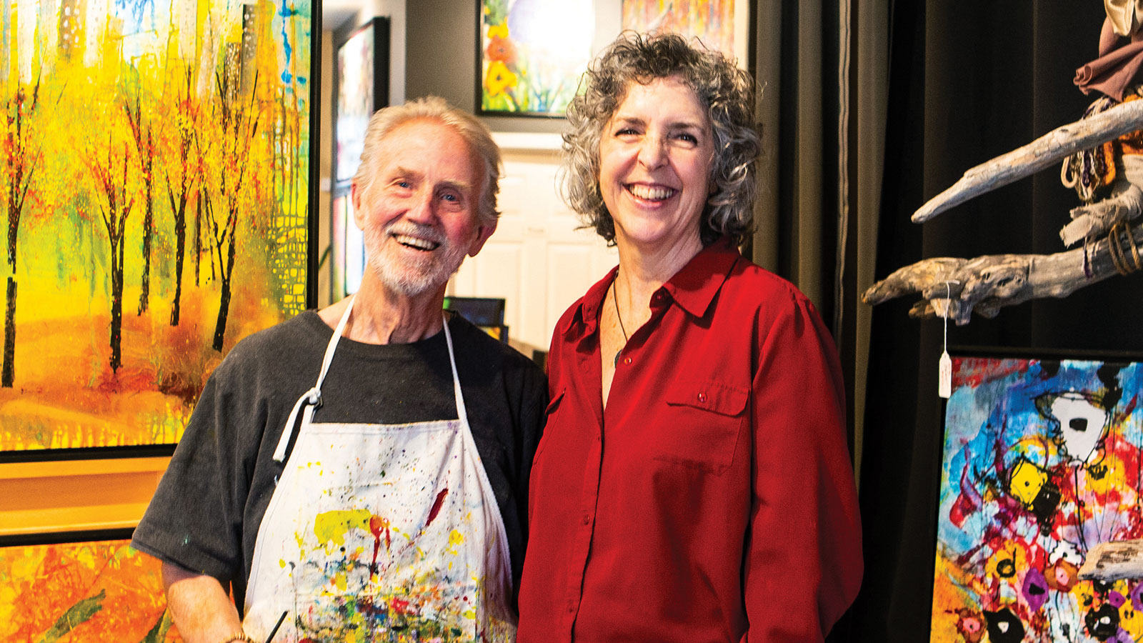 Man with painted apron and woman standing next to him, both in front of colorful paintings.