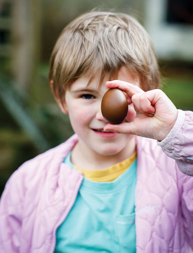 Girl in pink jacked holds chocolate-brown egg in front of her face