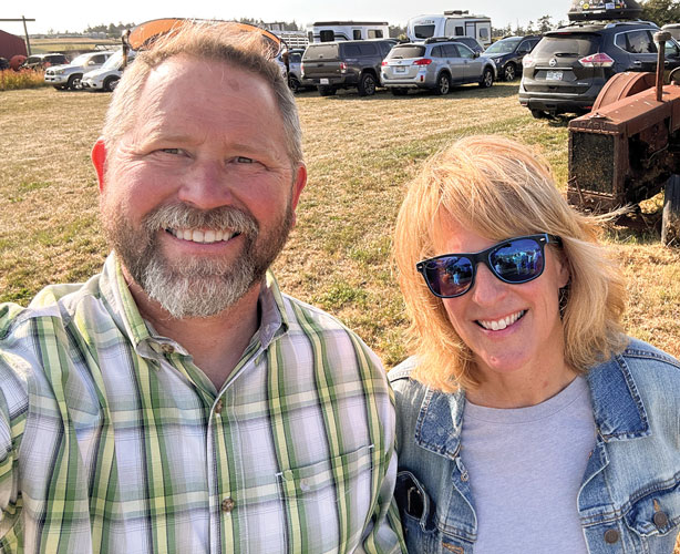 Man stands next to woman in sunglasses with cars parked on a grassy field in the background