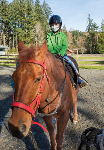 Girl wearing black helmet, green jacked and a surgical mask rides a brown horse