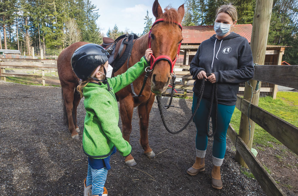 Young girl wearing a black helmet and green jacked pets brown horse while woman in grey sweatshirt looks on