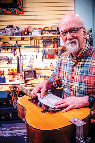 Older man in blue and red plaid shirt holds a tracing paper above an acoustic guitar