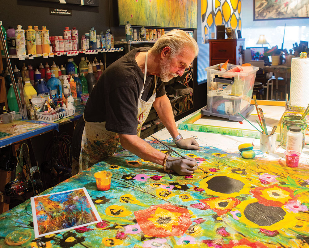 Older man with apron paints flowers on large canvas on a table
